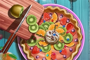 Ovocný koláč dekorovaný plátkami kiwi, pomaranča, jahodami, šľahačkou a farebnými guľôčkami na modrom stole, vedľa ktorého je doska s krájaným kiwi a nožom