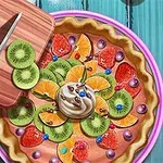Ovocný koláč dekorovaný plátkami kiwi, pomaranča, jahodami, šľahačkou a farebnými guľôčkami na modrom stole, vedľa ktorého je doska s krájaným kiwi a nožom
