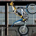 Motocyklista v žltočiernom oblečení prekonáva prekážku na motocykli pred industriálnym pozadím